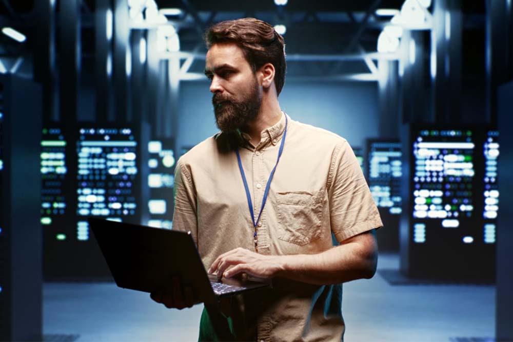Ein bärtiger IT-Expert mit Laptop steht in der Serverhalle und schaut nach links