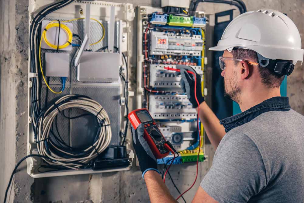 Ein Elektriker mit weißem Schutzhelm und dunklen Handschuhen kontrolliert eine Schalttafel mit einem Multimeter